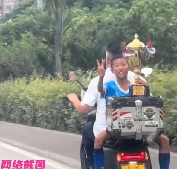 某个小男孩高举着手里的奖杯，还反坐在家长的电瓶车后座上，此刻小男孩把手里的奖杯放