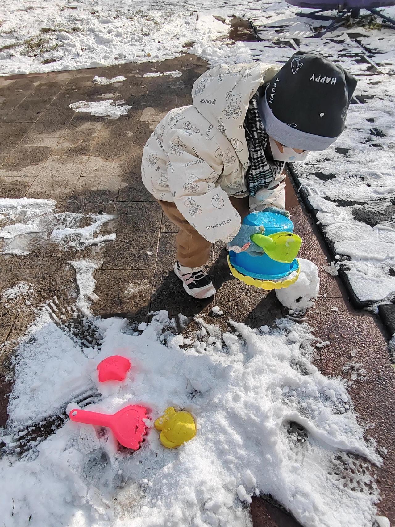 雪后太阳出来，小家伙裹着熊纹棉服蹲在雪边，彩铲“啪嗒”拍实桶里的雪，粉勺黄模半埋