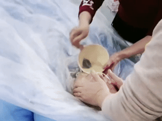 人老了真的很可怜，
一名护工摁住老人的头部，身上披着塑料膜，另一人一口一口往老人