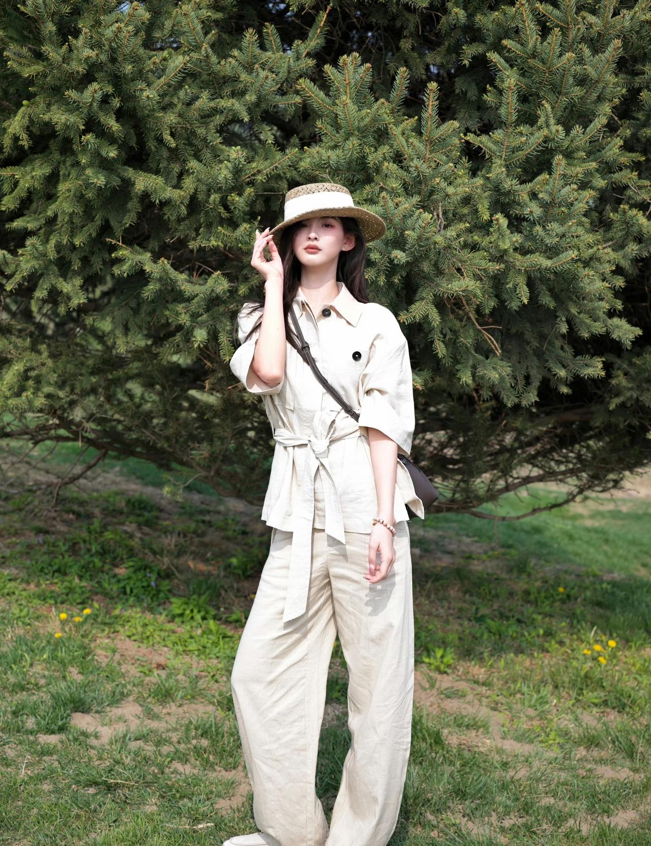 头戴草帽，身着米白色的短袖系带衬衫和同色系阔腿裤，脚穿白色鞋子，斜挎着包，姿态优