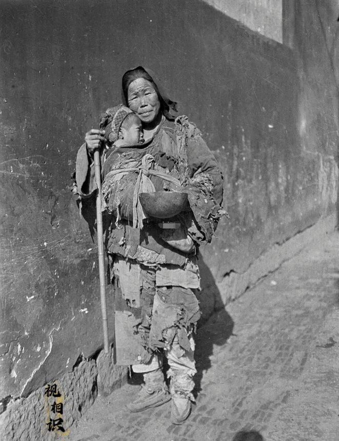1919年，浙江杭州，一对沿街乞讨的母子的照片，镜头中母亲衣衫褴褛，手中拿着一个...