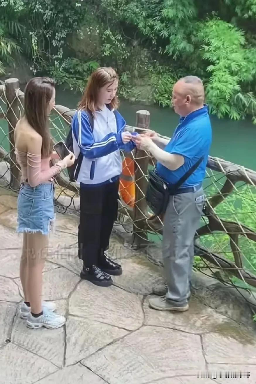 两个女孩在瀑布景区拍照，离她们不远处，有一位老人紧紧地盯着正在拍照的女孩看。女孩