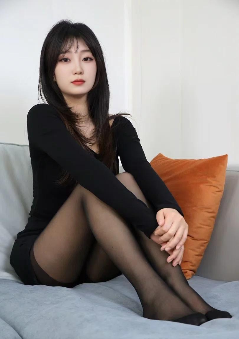 她穿着黑色修身连衣裙和黑丝袜，跪坐在浅灰色沙发上。背景是贴着软木板的白墙，整体氛