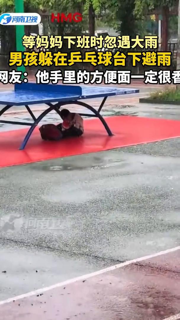 等妈妈下班时忽遇大雨，男孩躲在乒乓球台下避雨。网友：他手里的方便面一定很香。
河