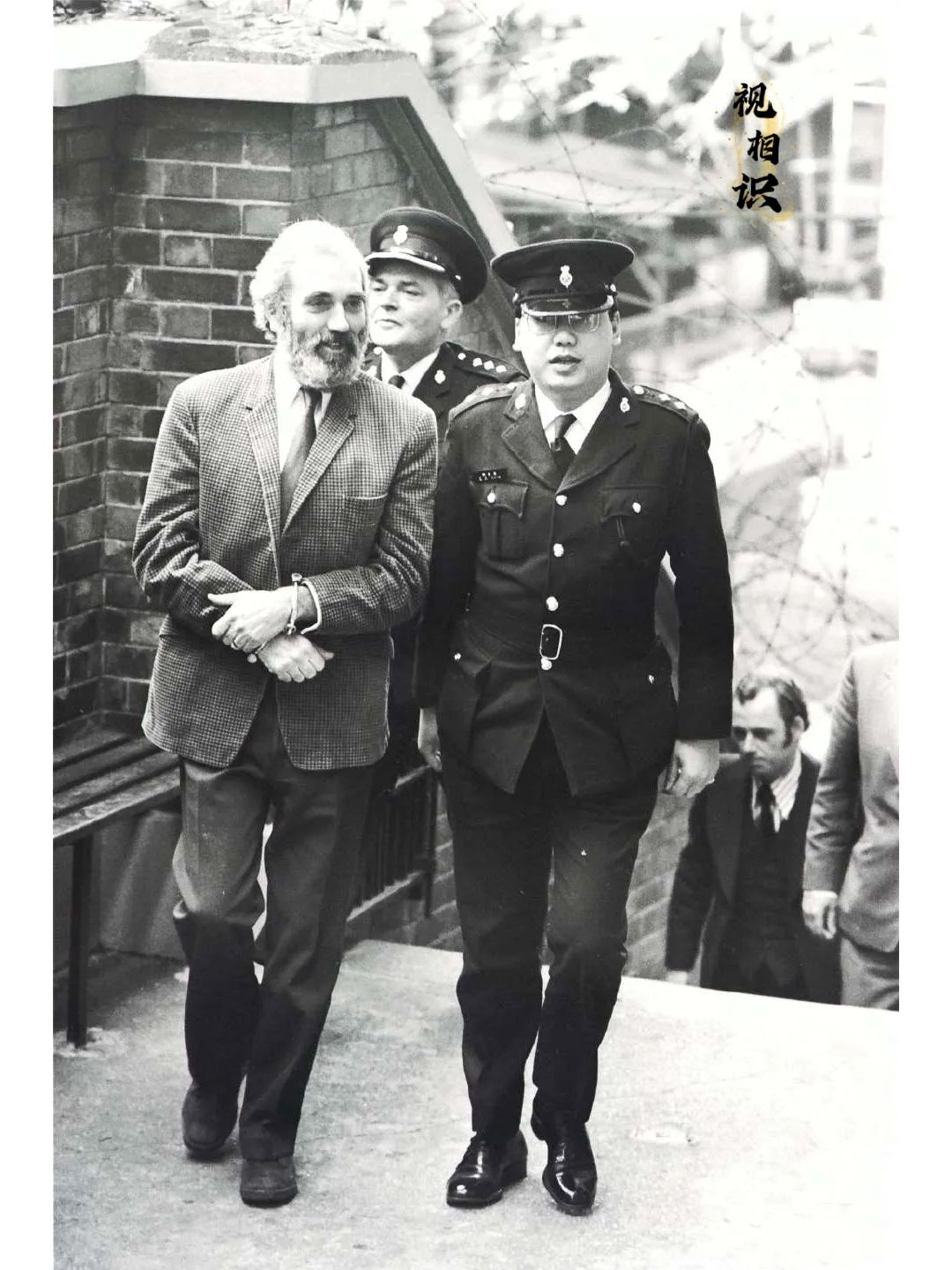 1975年，香港，前总警司葛柏在两名警察的陪同下，正步入法庭。他留着浓密的大胡子...