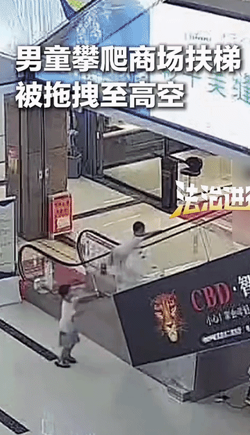 两名男孩在某商场的扶梯上玩闹。一名男孩攀爬时不慎被扶梯往上拖拽，另外一名男孩在旁