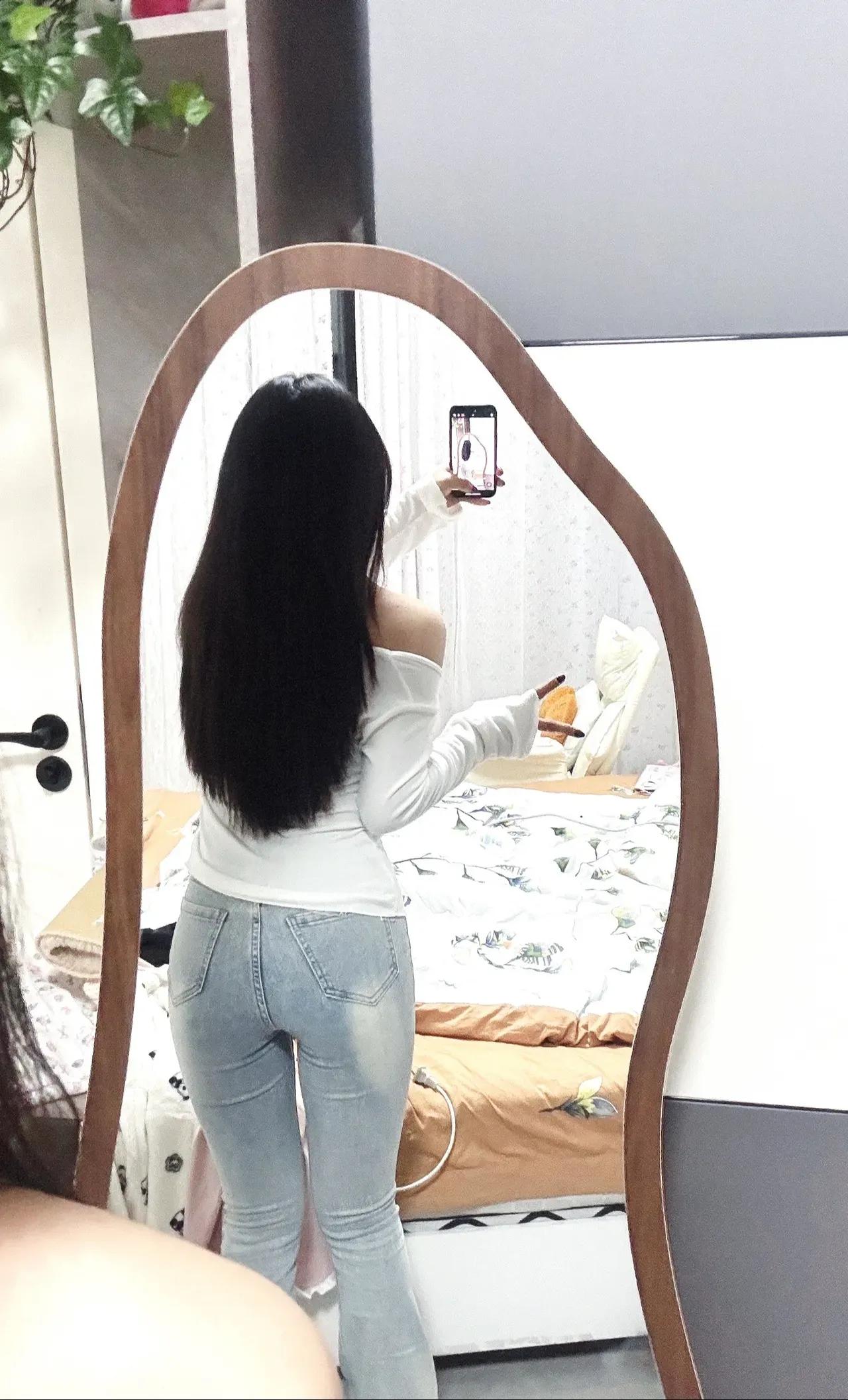 一位长发少女在镜子前自拍。她身穿黑白条纹短袖上衣和浅蓝色紧身牛仔裤，右手拿着一部