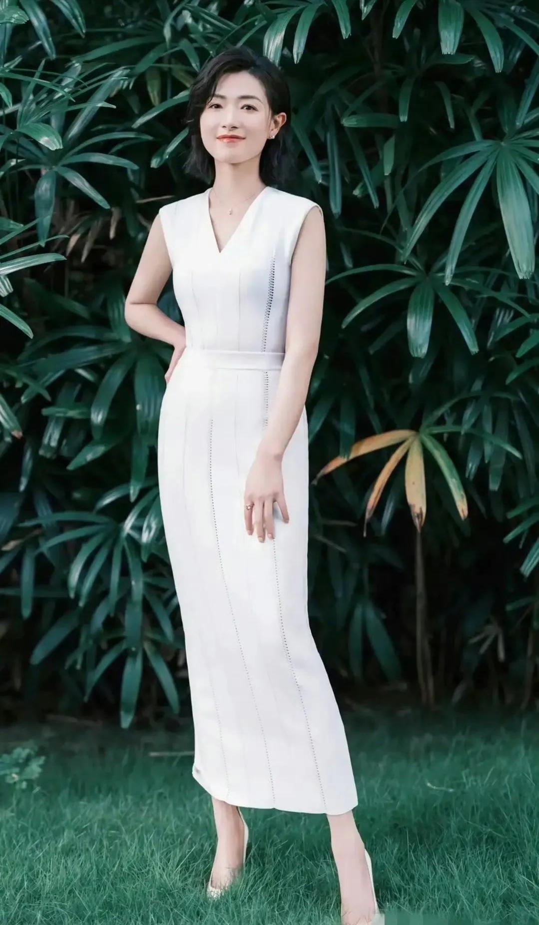 照片中的她，站在绿色的草坪上。一身白色束身长裙。搭配着一双高跟鞋。让她本就苗条的