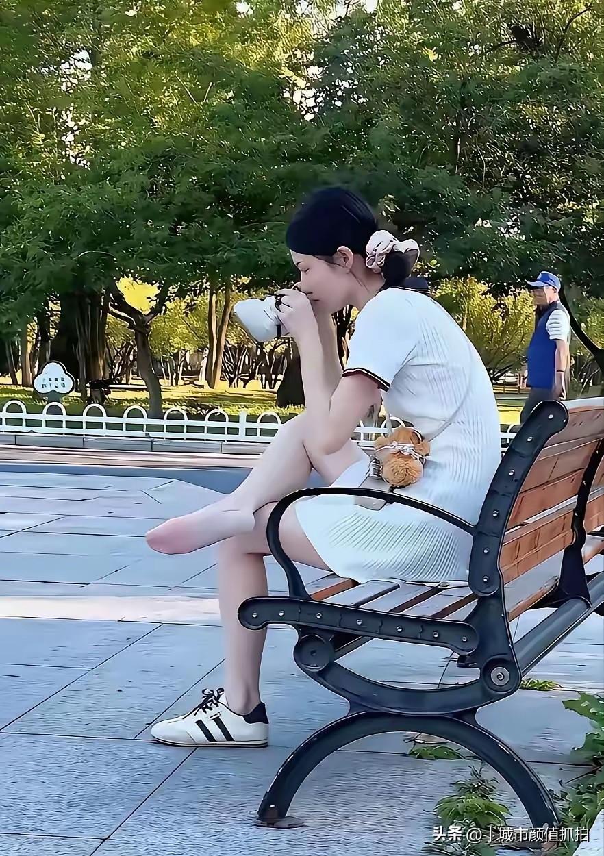 秋日午后的公园浸在暖融融的金色光影里，姑娘身着简约白针织裙，扎着松散发髻，坐在木