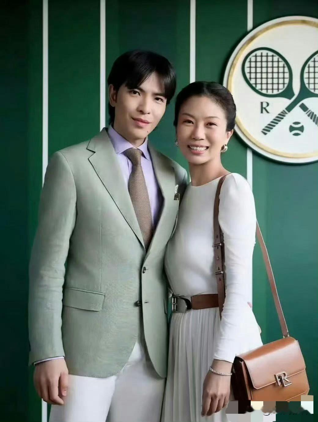 这是歌手萧敬腾与妻子林有慧一同现身温布尔登网球锦标赛（温网）现场的活动合影。画面中，37岁的萧敬腾身着浅绿色西装搭配白裤，显得儒雅干练；54岁的林有慧则身穿白色长袖连衣裙配棕色腰带及同色系单肩包，造型简约优雅。两人紧靠站立面对镜头微笑，背景为标志性的深绿色竖条纹墙面及温网Logo，整体着装正式讲究，