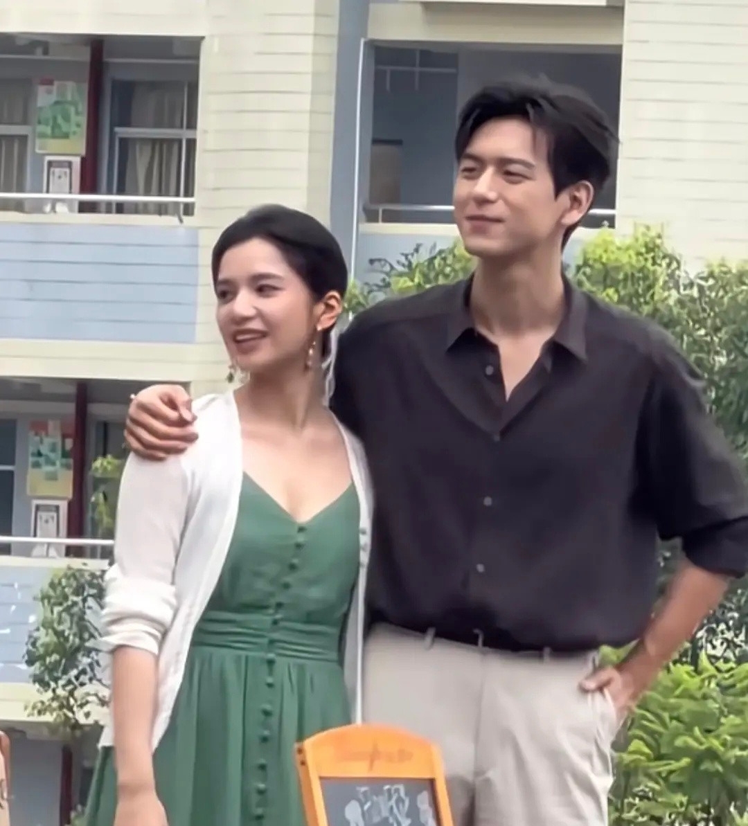 剧组户外路透流出，二人同框画面十分养眼。张婧仪一身墨绿色吊带长裙，外搭一件柔和白开衫，温婉复古，气质柔和恬淡。李现身穿松弛黑衬衫搭配米白休闲西裤，单手插兜，成熟儒雅的年代男主感扑面而来。
 
男生手臂轻搭女生肩头，姿态自然松弛。前景小黑板道具加持，浓浓的年代故事感扑面而来。生图状态在线，二人气质适配