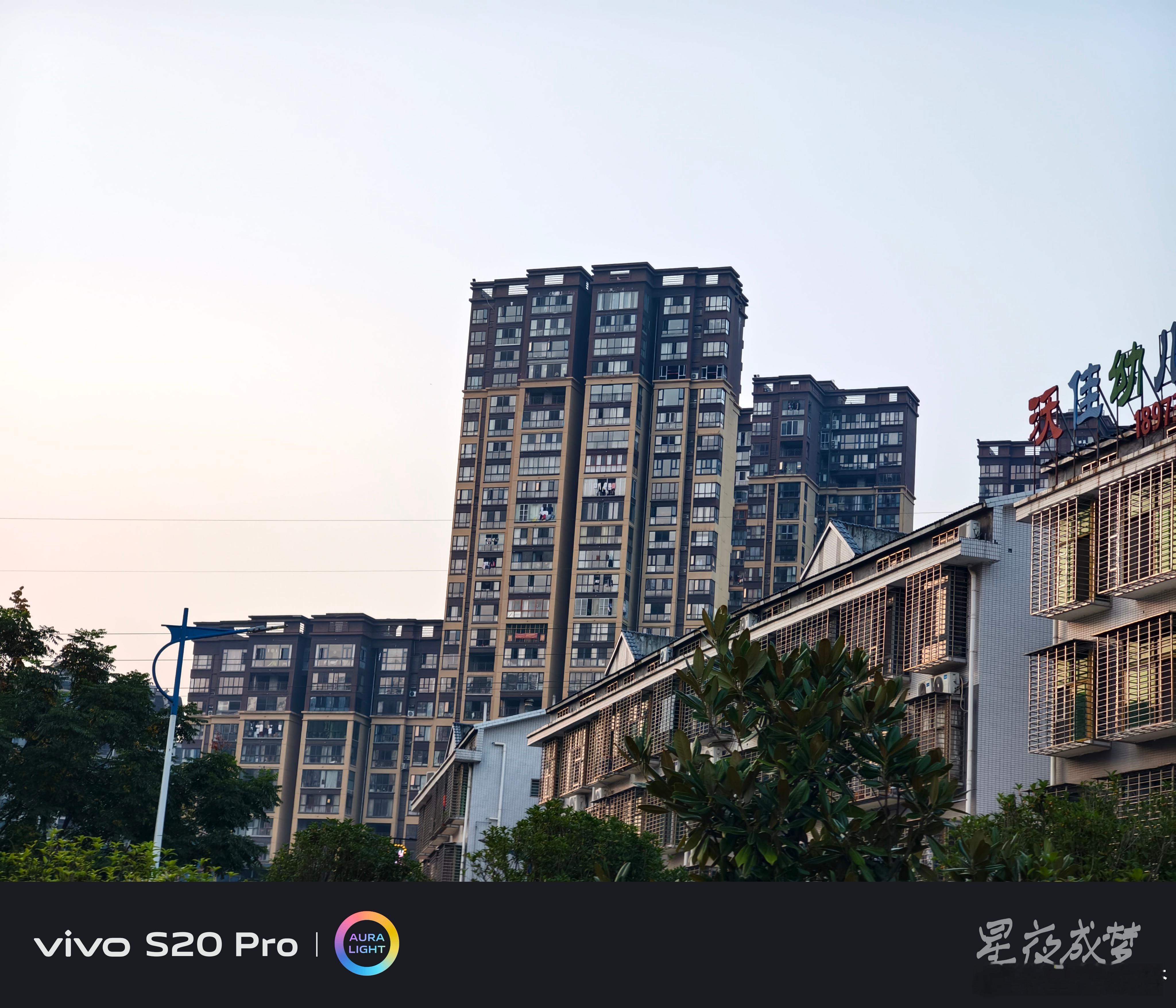 #vivo[超话]# 仰起头看这几栋高层住宅，棕色和米色的外墙在黄昏的光线下很温暖，窗户排列整齐，有的阳台上还晾着衣服，充满了生活气息，旁边的低矮建筑上"沃佳幼儿园"的招牌清晰可见，绿树在下方点缀。vivo S20 Pro的解析力在线，建筑的细节和窗户的纹理都清晰锐利，仰拍的透视感很到位，随手记录城市 ​