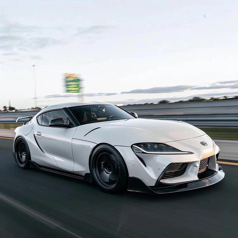 白色丰田Supra，黑化套件加身，机舱暗藏玄机
一台白色的丰田Supra，配上黑色的空力套件，走到哪里都是视觉中心。前脸大包围加上风刀，看着挺有攻击性。车尾的鸭尾扰流板配合底下的三角形领航灯，排气声浪先不说，视觉压迫感已经拉满了。侧面深色轮毂里透出红色刹车，车身姿态压得非常低，紧贴地面。打开发动机盖