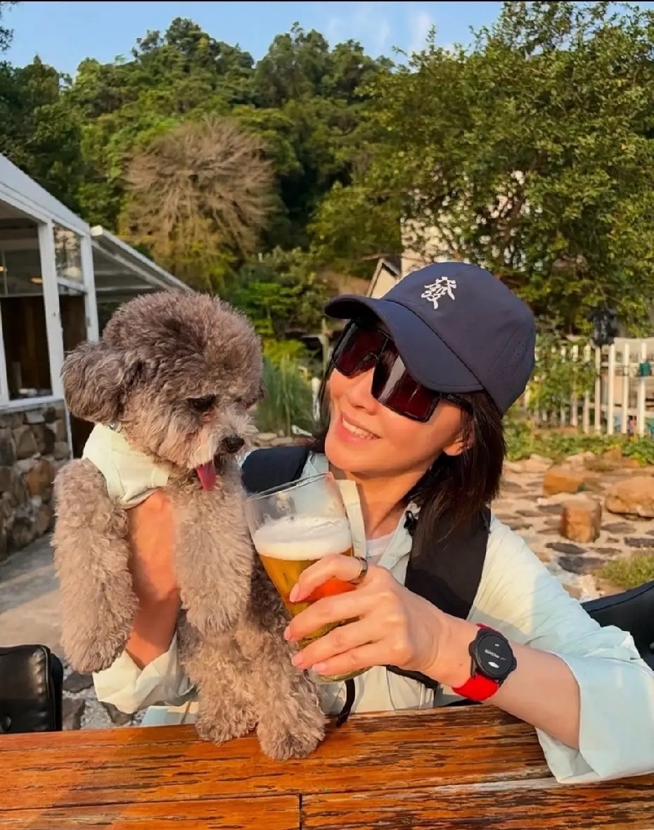 60发刘嘉玲分享她的灰色泰迪爱犬户外休闲合影，她头戴印有“發”字的深色棒球帽，佩戴大框墨镜，身着浅色系户外休闲上衣，状态松弛愉悦。她手中举着一杯带有绵密泡沫的饮品，正和怀里的小狗互动，仿佛要和爱犬“碰杯”，画面充满生活趣味。
如今却满心宠溺地照料家中三只爱犬，除了这只灰色小泰迪，还有一黑一白两只拉布