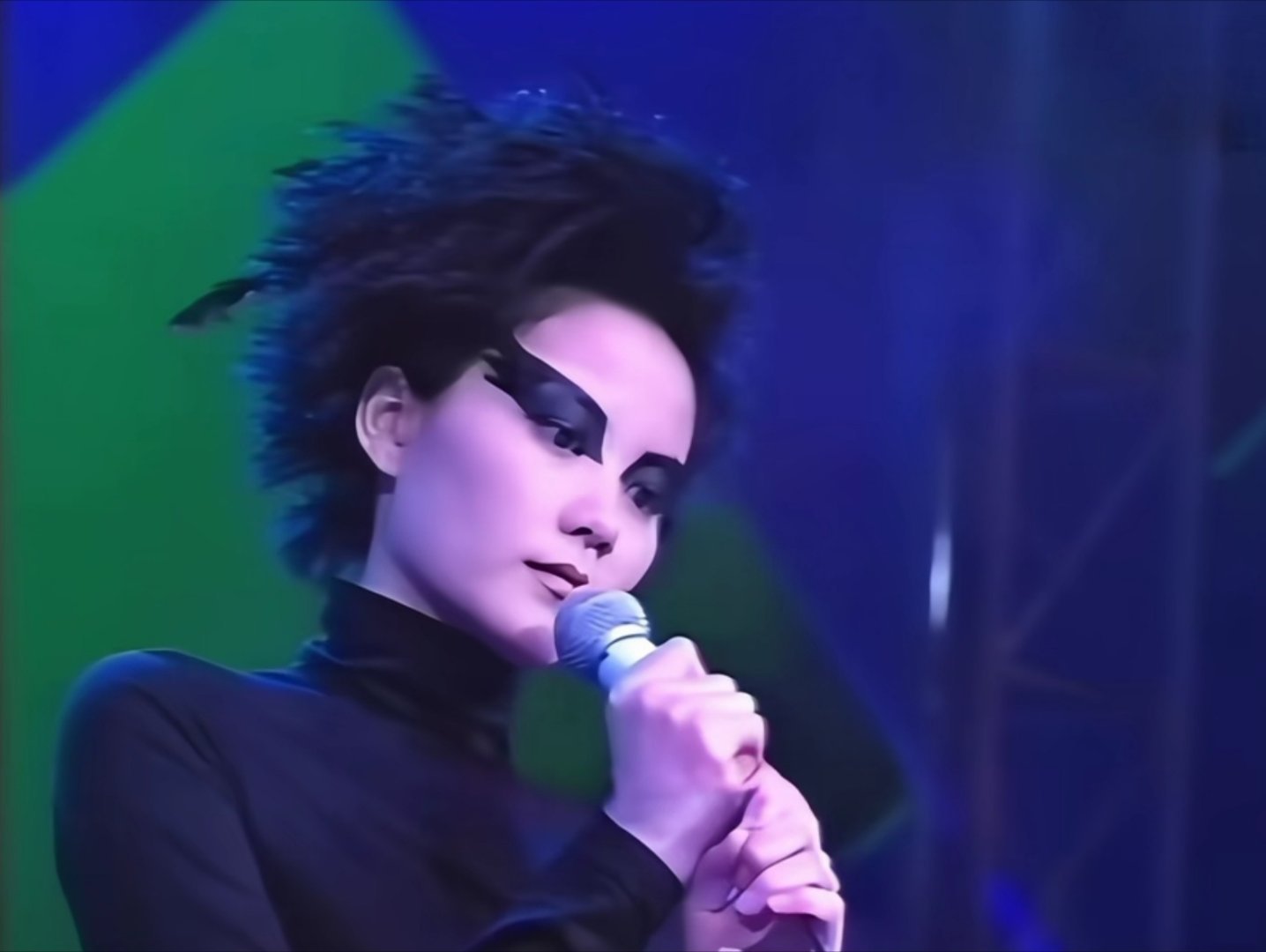 王菲97年演唱《只愿为你守着约》的妆造

纯黑覆盖式眼妆向颧骨外铺展开，眼型被拉得又尖又长，像猛禽羽翼、也像晕开的暗影，黑发蓬松凌乱、黑衣黑裤纤细挺括，多了鬼魅感，一双杏眼凝望时好似隔了一层雾气，后面的交响乐是幽冥之间流动的回响，黑青色和苹绿色的背景漫无边际地糅杂在一起，是女妖转世对 ​