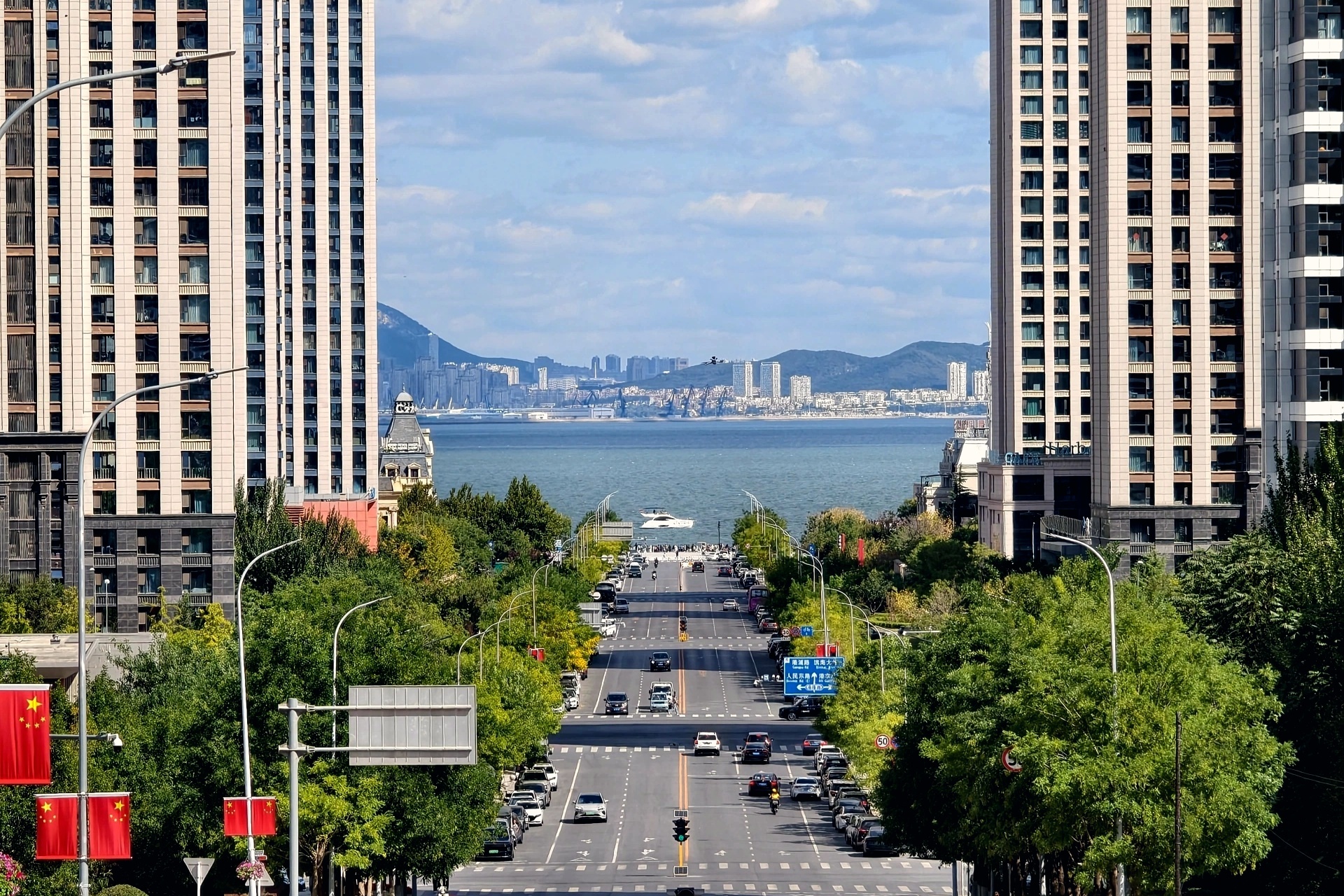 大连港东五街｜奔赴一条通向大海的路 东北超燃AI看大连盛世华章悦享大连万千气象看辽宁 来大连东港，一定要打卡港东五街。这条火爆全网的网红街道，是独属于大连的浪漫，一条笔直缓坡大道，向着蔚蓝海面无限延伸，一眼望穿楼宇与碧海，自带电影氛围感。全长1.5公里的街道南高北低，站在坡顶远眺，两侧现代高楼天然形成画框，框住远方无垠大海。最动人的瞬间，便是等候巨轮穿楼而过。每日上午10:50、下午16:30左右，客轮伴着悠长鸣笛，缓缓从楼宇缝隙驶出，白船、蓝天、碧海同框，定格震撼治愈的画面，无数摄影爱好者专程前来蹲守抓拍。不必执着于等船，漫步街道本身就是享受。干净开阔的路面，搭配通透的海天背景，长焦随手一拍，就是氛围感满满的人像大片。沿着道路一直向北前行，便可直达海边木栈道，海风扑面而来，远眺港口船吊与海岛，城市摩登质感与海洋的辽阔在此相融。运气好时，还能偶遇英姿飒爽的女骑警巡街，成为意外的惊喜。这里与威尼斯水城近在咫尺，打卡结束可以顺路漫步水巷，一站式感受东港的多重浪漫。无需门票，全天开放，地...