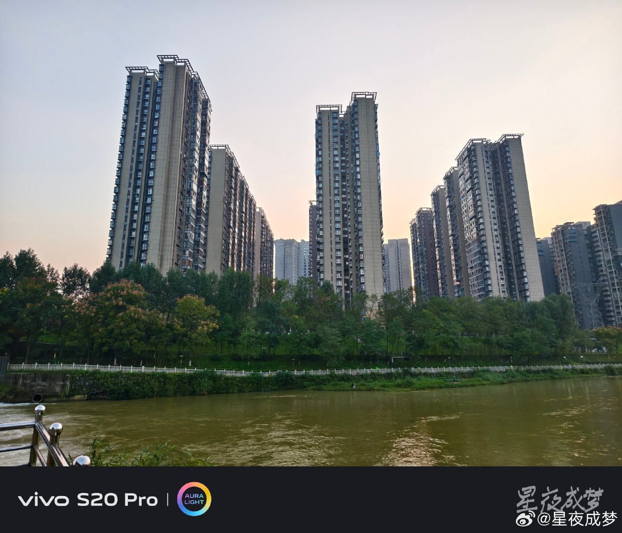 #vivo[超话]# 黄昏时分的河边太美了，几栋高层住宅楼在暖橙色的天空下格外醒目，河水在下方静静流淌，岸边的绿树成荫，白色的栏杆沿着河岸延伸，城市的傍晚温柔又治愈。用vivo S20 Pro记录下这一刻，AURA LIGHT把天空的暖色调和建筑的轮廓都还原得很到位，画面通透有质感。#vivo##vivo超话# ​