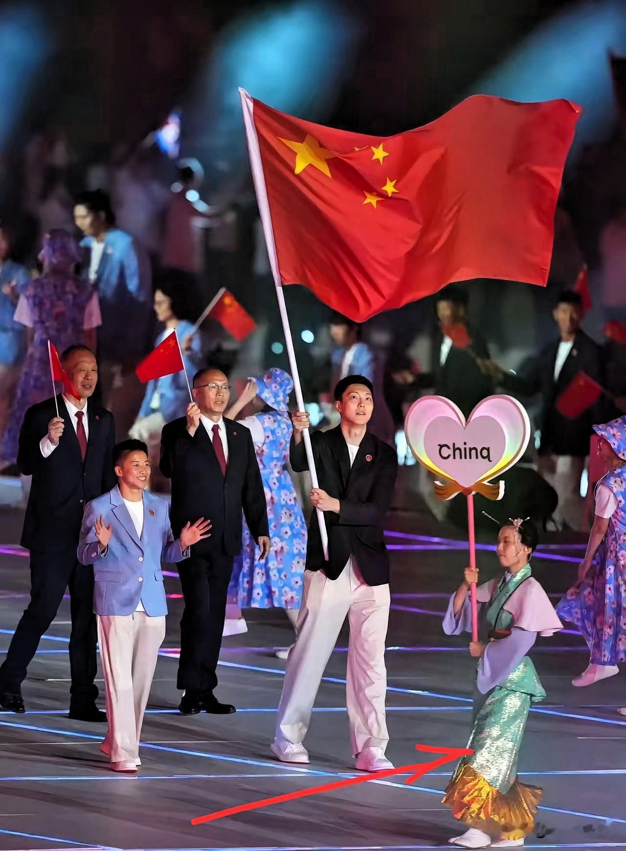 亚运会开幕式上，中国代表团入场那一刻，旗手气场全开，五星红旗猎猎生风，看得人热血直冲头顶。可镜头一扫到旁边举爱心牌的引导员，画风瞬间变了——这套造型，真让人一时语塞。

先说这身穿搭，配色和面料凑在一起，怎么看怎么别扭，第一眼就透着股说不出的违和感。再瞧妆容、神态和动作，不少网友直呼看得脚趾抠地，恨