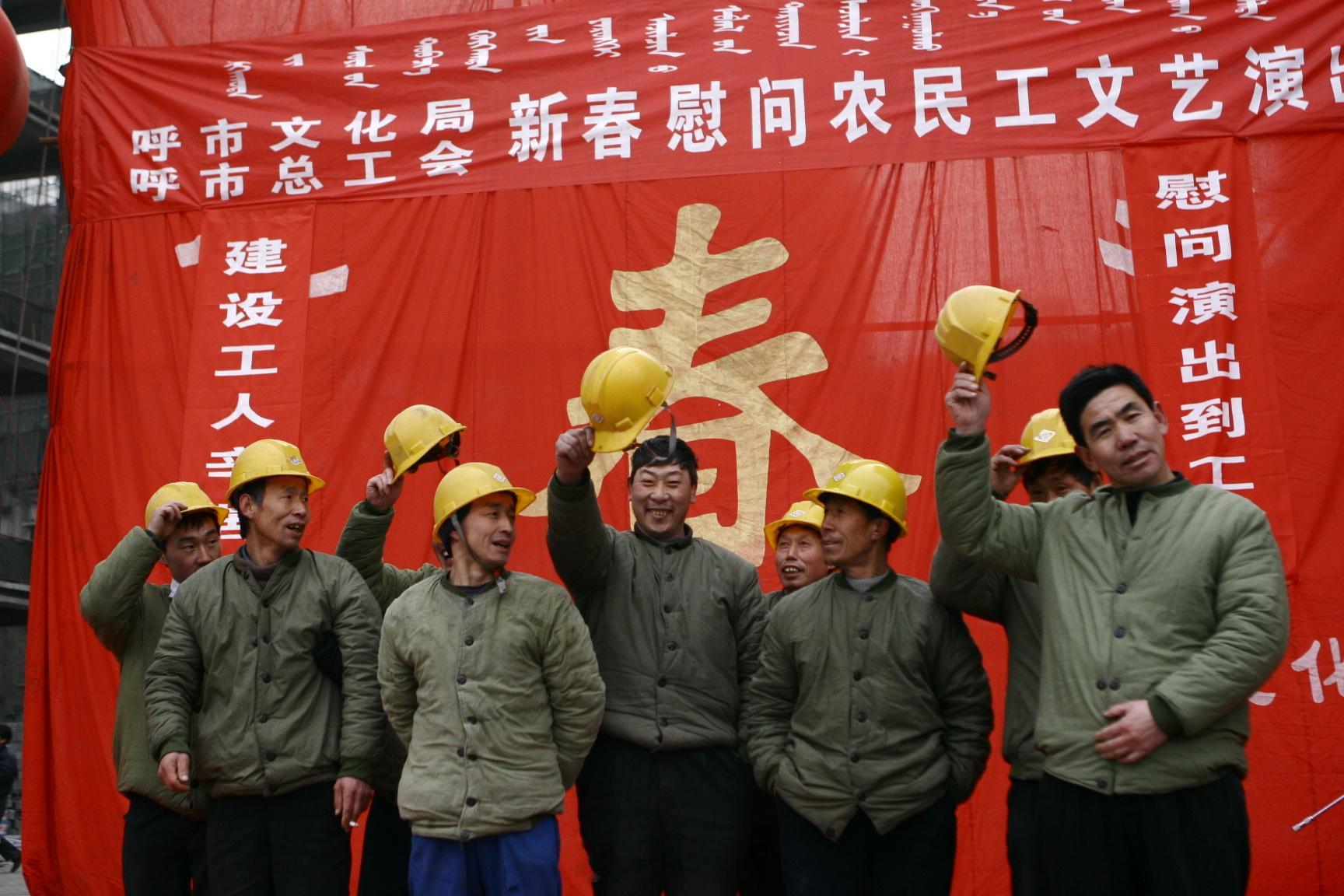 #呼和浩特老照片# 红底背景布前，一群穿绿工装的农民工站得整整齐齐，有的举着黄色安全帽，有的正扶正帽沿，脸上都是笑。背景上金灿灿的"春"字和蒙文标语格外醒目，"呼市总工会""文化局"的横幅写着"新春慰问农民工文艺演出"。那年头的年根底下，慰问演出队下工地，演员在台上唱，工人在台下看，工地上 ​