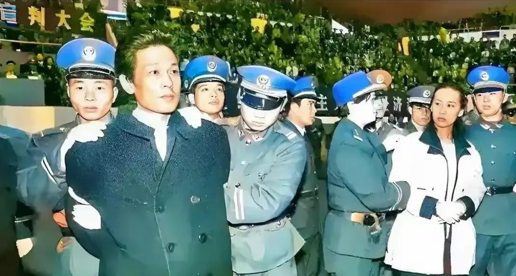 这张照片拍摄于1995年12月28日，记录了深圳“王建业、史燕青贪腐案”公审宣判大会的现场。

图中被法警押解的男子是原深圳市计划局财贸处处长王建业，他因受贿罪被判处死刑；身旁的女子是他的重婚妻子史燕青，因投机倒把罪、贪污罪被判处死缓。

王建业在1992年至1993年间，利用审批进口物资指标和外汇