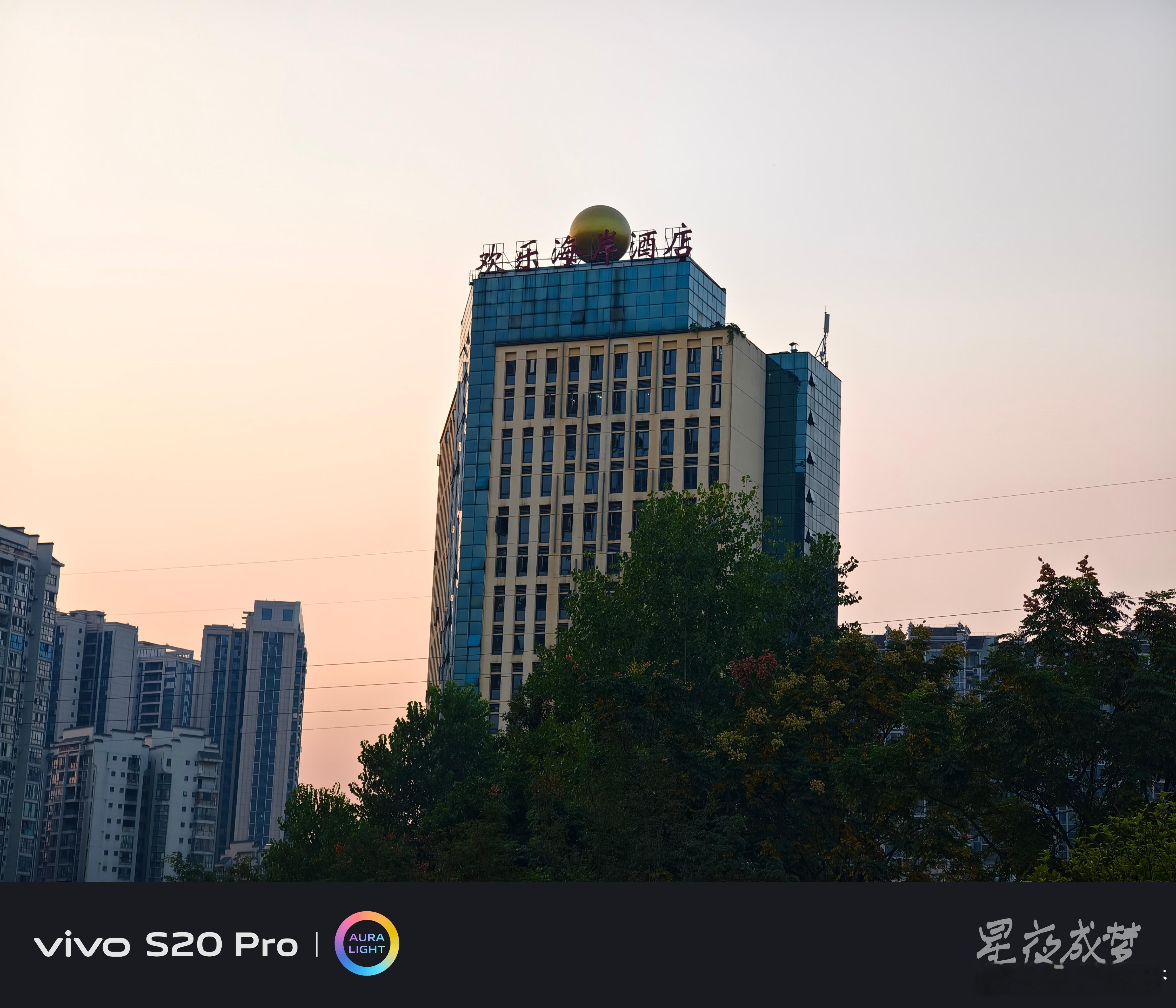 #vivo[超话]# 这栋"欢乐海岸酒店"太有辨识度了，蓝色的玻璃幕墙在黄昏的天空下格外醒目，顶部的金色圆球和红色大字招牌很有特色，周围的绿树环绕着建筑，暖橙色的天空做背景，城市地标感十足。用vivo S20 Pro仰拍定格，建筑的线条笔直锐利，玻璃幕墙的反光清晰可见，色彩通透，随手拍就是好照片。#vivo ​