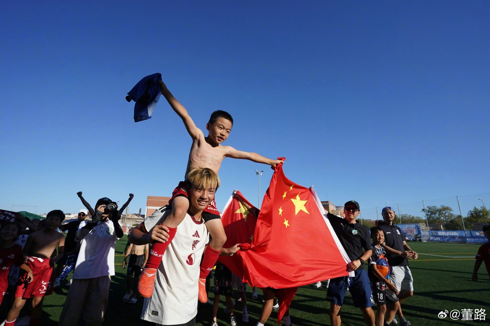 李大哥肩膀托举起qiao大哥……（智能识图）：这是“中国足球小将”在西班牙捧杯夺冠后的庆祝现场，画面中一名身穿白色球衣的球员将一名赤膊小队员扛在肩头，少年高举一面鲜艳的五星红旗和一件蓝色衣物，神情兴奋且充满自豪。这张照片由知名足球评论人董路发布，记录了该青少年足球队在海外赛事中战胜对 ​
