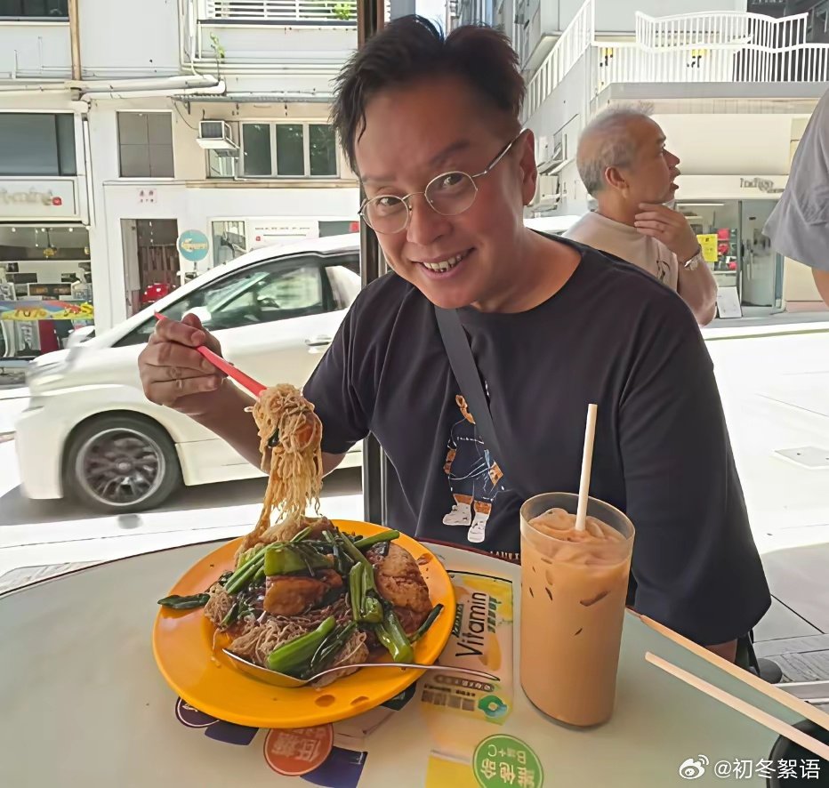谭咏麟坐在香港街边露天小吃摊前座位上，吃着一份炒米粉，喝着一杯冻奶茶，没有隆重的排场，一身休闲T恤，简简单单的享受，非常的惬意。 ​